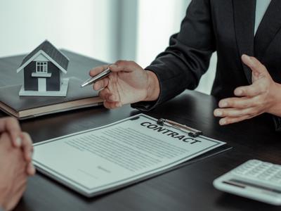 Two people discussing a real estate contract, with a small model house on the table and the contract document prominently displayed.