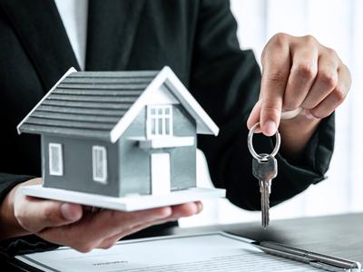 A person in a suit holding a small model house in one hand and a set of keys in the other, symbolizing real estate transactions.