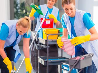 Three cleaning staff members wearing blue uniforms and yellow gloves work with cleaning supplies and equipment inside a bright indoor space.