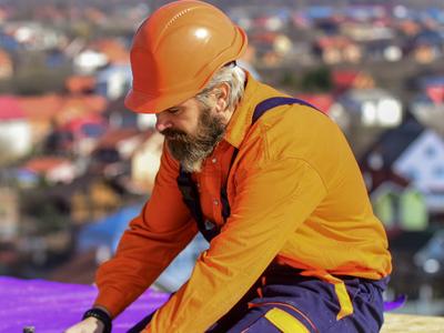 A bearded construction worker wearing an orange hard hat and orange outfit, working on a roof with a residential neighborhood in the background.