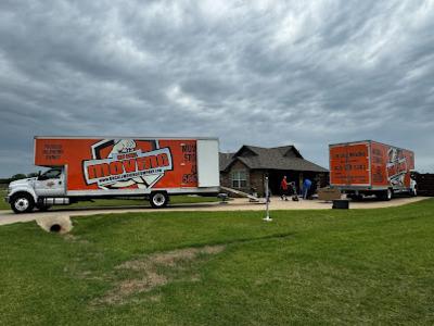 Two large orange trucks from a moving company were parked outside a suburban home. Movers were working near the entrance under a cloudy sky.