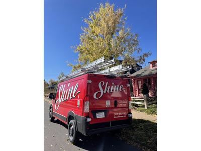 A red Shine van parked on the side of the road, equipped with ladders on the roof, likely used for window cleaning or gutter services. The vehicle is branded with the Shine logo in large white script.
