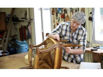 The image shows an artisan meticulously working on an antique chair in a workshop, focused on restoring or reupholstering its frame. The setting is a traditional workshop with tools visible in the background, indicating a hands-on approach to craftsmanshi