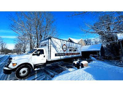 A moving truck parked in a residential neighborhood during wintertime. The truck is branded with the logo "Firefighters Moving Co." and appears to be positioned in a driveway with snow covering the ground and surrounding trees. The clear blue sky contra