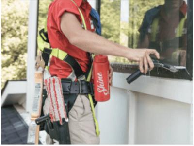 Professional window cleaner in a red uniform cleaning an exterior window using a squeegee, with cleaning tools and a 'Shine' branded water bottle strapped to his belt.