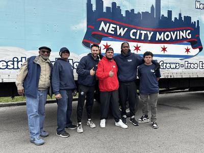 The image shows a group of movers from New City Moving posing in front of their moving truck, giving thumbs up. The truck features the company's logo, which includes a city skyline and red stars, emphasizing their Chicago-based moving services.