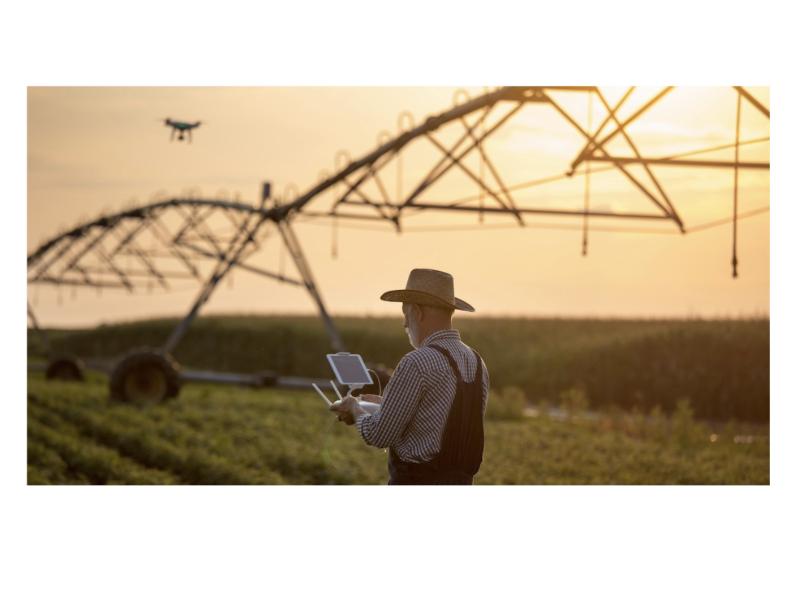 This image shows a farmer in a field at sunset operating a drone using a remote controller or tablet. The background includes an agricultural irrigation system, symbolizing the integration of technology in modern farming practices.