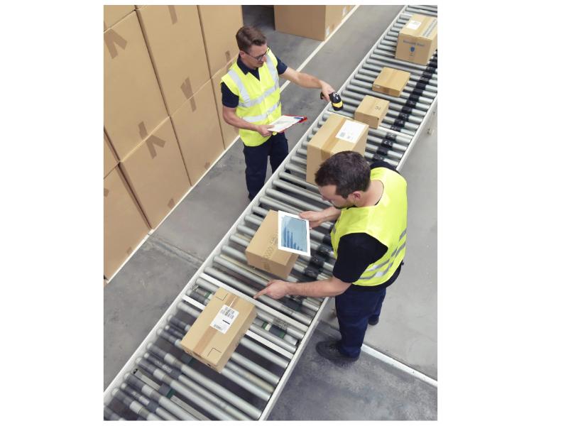 Two warehouse workers in high-visibility vests scan and inspect packages on a conveyor belt, ensuring efficient processing and shipment.