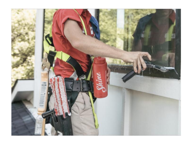 A professional window cleaner uses a squeegee on a large glass surface, equipped with tools and safety gear for the job.