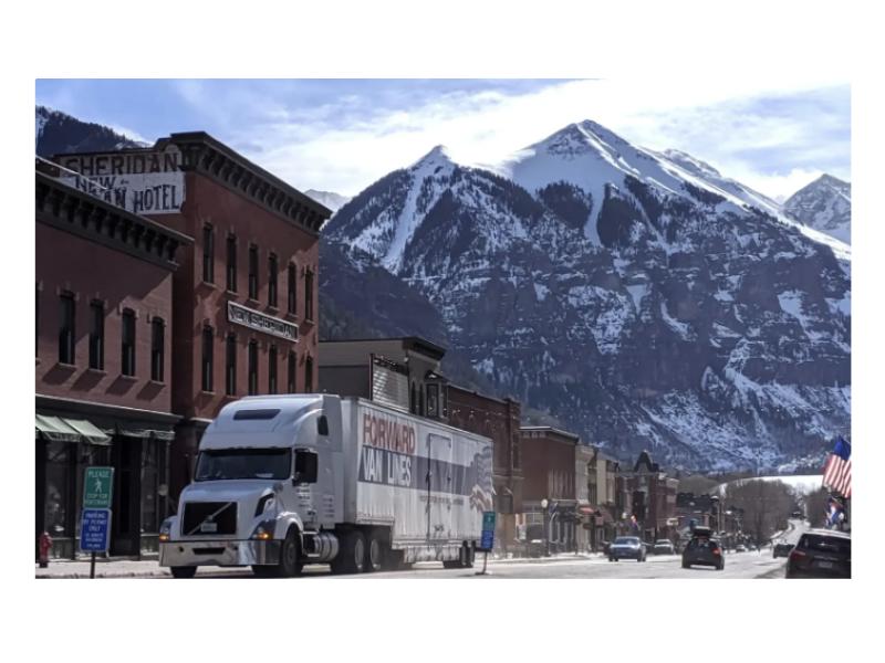 A large Forward Van Lines truck drives through a scenic mountain town with snow-covered peaks in the background.