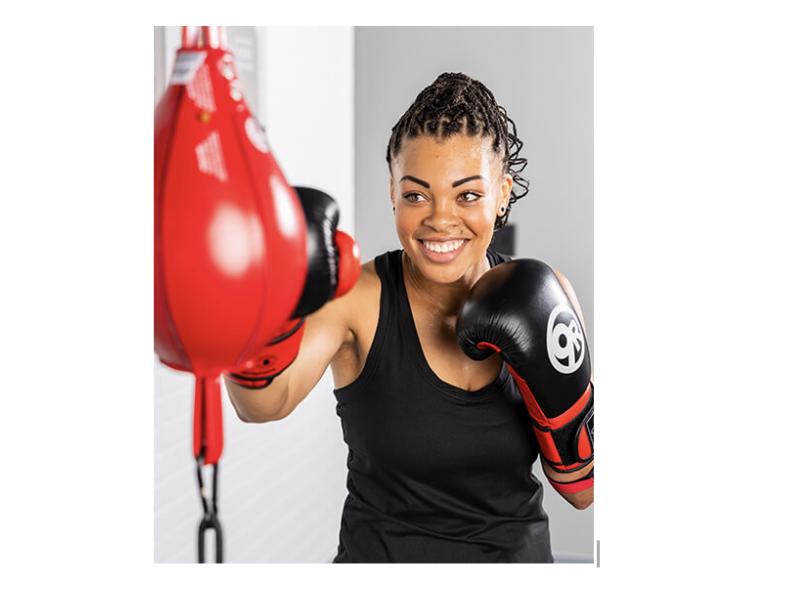 The image shows a woman in boxing gloves smiling as she trains with a speed bag, highlighting her focus and enjoyment during a workout session.