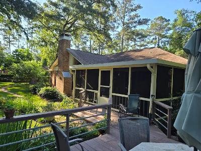 The image captures a charming, sunlit rear exterior of a single-story home with a large screened-in porch and an adjoining outdoor deck. The area is surrounded by lush green foliage and mature trees, creating a private, backyard oasis. This type of home r