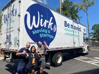 A large white moving truck with a striking dark blue vinyl wrap featuring the Wirks Moving & Storage logo parked on a quiet residential street in front of a substantial house. The second photo (unnamed_imresizer.jpg) shows three smiling people--two crew m