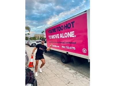 The image shows a smiling mover, wearing a black company t-shirt and shorts, posing in front of a bright pink moving truck with bold white text that reads, "YOU'RE TOO HOT TO MOVE ALONE." The truck clearly advertises Flexdolly Moving & Delivery with th
