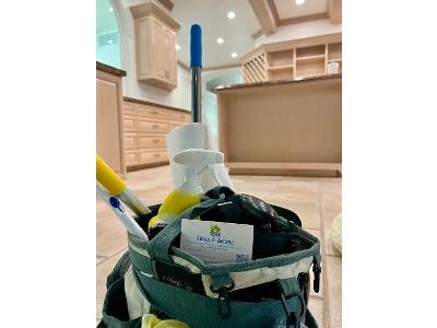 The caddy is filled with cleaning essentials, including a blue-handled duster or mop, a roll of paper towels, various spray and squeeze bottles, and a business card for Clean & Serene Dayton, LLC is prominently displayed on the front of the bag. The backg