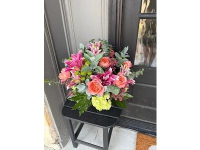 The image captures a beautiful, vibrant floral arrangement resting on a small, dark stool outside a home entrance.