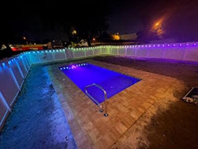 The photograph shows a freshly constructed inground pool surrounded by light-colored travertine or paver decking. The water is glowing a deep blue from the interior lighting. A safety fence surrounds the area, and what makes the scene unique is the multi-