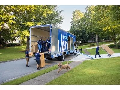 Captures a sunny, professional moving scene where a Wirks Moving & Storage crew is actively loading a large, bright blue-and-white truck on a residential street. Multiple smiling, uniformed movers work efficiently to carry boxes and use a ramp to load a d