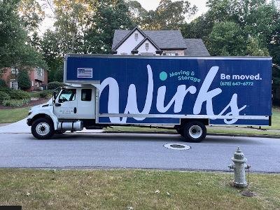 A large white moving truck with a striking dark blue vinyl wrap featuring the Wirks Moving & Storage logo is parked in front of a residential home on a quiet, tree-lined street. The phone number (678) 647-6772 and the tagline "Be moved." are clearly vis