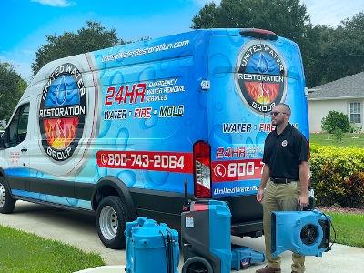 A uniformed technician from United Water Restoration Group stands ready in a residential driveway with specialized drying equipment, next to a large blue and red van advertising 24HR EMERGENCY services for WATER . FIRE . MOLD restoration.