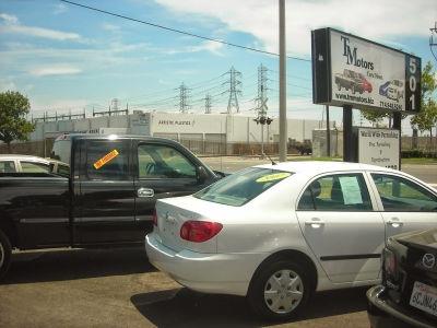The exterior of the TM Motors dealership features a white sedan and a black pickup truck parked in the lot. A large business sign displays the company name and website, set against a backdrop of industrial power lines and a bright sky.